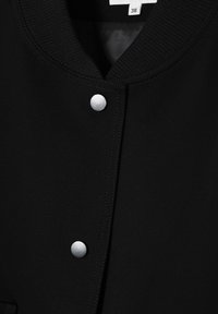 Černá látková bunda s žebrovaným límcem, s stříbrnými druky a viditelným prošíváním podél okrajů. Velikostní štítek je 38.