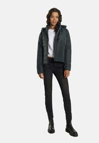 Mörkgrön puffjacka med huva, quiltat mönster och framzipper. Bärs över en vit stickad topp, i kombination med svarta jeans och ankelstövlar.