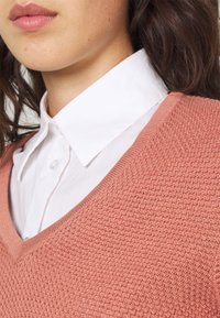 Pfirsichfarbener, strukturierter Strickpullover mit V-Ausschnitt, über einem weißen Hemd mit Kragen getragen. Der Stoff wirkt weich und atmungsaktiv.