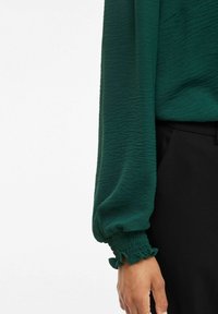 Närbild av en persons arm som bär en strukturerad mörkgrön långärmad blus med veckade elastiska manschetter och svarta byxor.