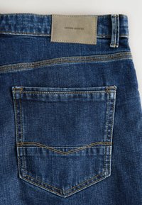 Blugi din denim albastru închis, cu un buzunar pe spate cu detaliu de cusătură tip chevron și o etichetă din material gri deschis embosată cu "DENIM SUPPLY."