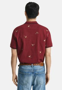 Mann in einem kastanienbraunen, kurzärmligen Poloshirt mit Vogelmotiven, in hellblauen Jeans gesteckt, mit einem geflochtenen braunen Ledergürtel, von hinten betrachtet.