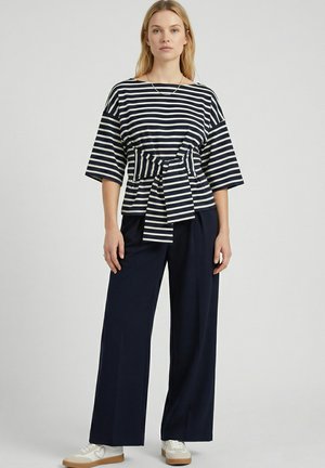Frau steht, trägt ein marineblau-weiß gestreiftes Oberteil mit gebundener Taille und weite marineblaue Hosen mit weißen Turnschuhen.