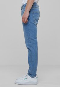 Jeans de mezclilla azul claro mostrados de lado, con un corte ajustado, diseño de cinco bolsillos y costuras oscuras. Llevados con zapatillas blancas.