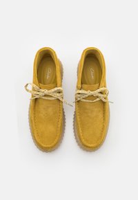 Ein Paar senfgelbe Wildlederschuhe für die Freizeit mit weißen und gelben Schnürsenkeln sowie dicken, geriffelten Gummisohlen, von oben gezeigt.