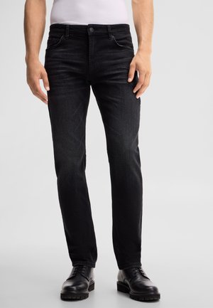 Mann trägt schmale schwarze Jeans mit schwarzen Leder-Herrenschuhen und steht vor einem schlichten Hintergrund.