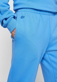 Person wearing bright blue sweatpants with elastic waistband and a small crocodile logo, hand in left pocket.