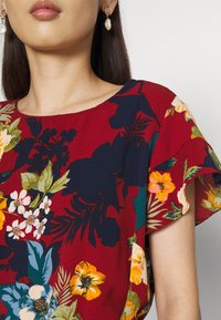 Röd blommig blus med korta, volangärmar. Har blommor i gult, vitt och blått mot en rik röd bakgrund. Mjuk tygkvalitet.