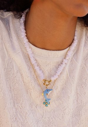 Nahaufnahme eines Halses, der ein weißes, strukturiertes Oberteil trägt, mit zwei Halsketten: einer groben weißen Perlenkette und einer goldenen Kette mit blauem Delfinanhänger.