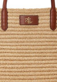 Geweven stro tote tas met bruine leren handvatten en een gouden logo plaatje. De tas heeft een gestructureerd, natuurlijk vezelontwerp met horizontale lijnen.