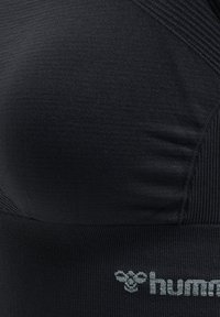 Zwarte sportbeha met een gladde textuur, voorzien van een V-vormig ontwerp, subtiele geribbelde accenten en een klein grijs logo aan de onderkant.
