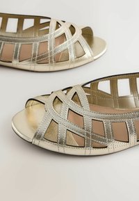 Ballerines métalliques dorées à bout ouvert avec un design de brides croisées et des découpes géométriques, exposées sur une surface claire.