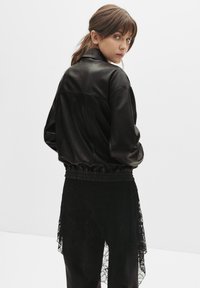 Čierna umelá kožená bomber bunda, oversized strih, elastický spodný lem a manžety. Model stojí s otočeným chrbtom, nosí vrstvené čierne čipkované spodky.