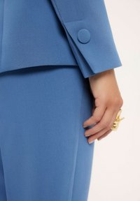 Niebieski dopasowany garnitur z teksturowanej tkaniny; posiada klapę z wycięciem oraz okrągły guzik na rękawie. Złoty pierścień na dłoni.
