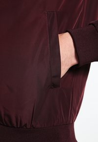 Burgunderfarbene Jacke mit glatter, glänzender Textur, gerippten Bündchen und einer Seitentasche. Der elastische Saum sorgt für eine figurbetonte Optik.