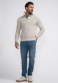 Pull en maille beige clair avec un col zippé, deux boutons et des poignets côtelés. Assorti avec un jean en denim bleu et des baskets blanches.