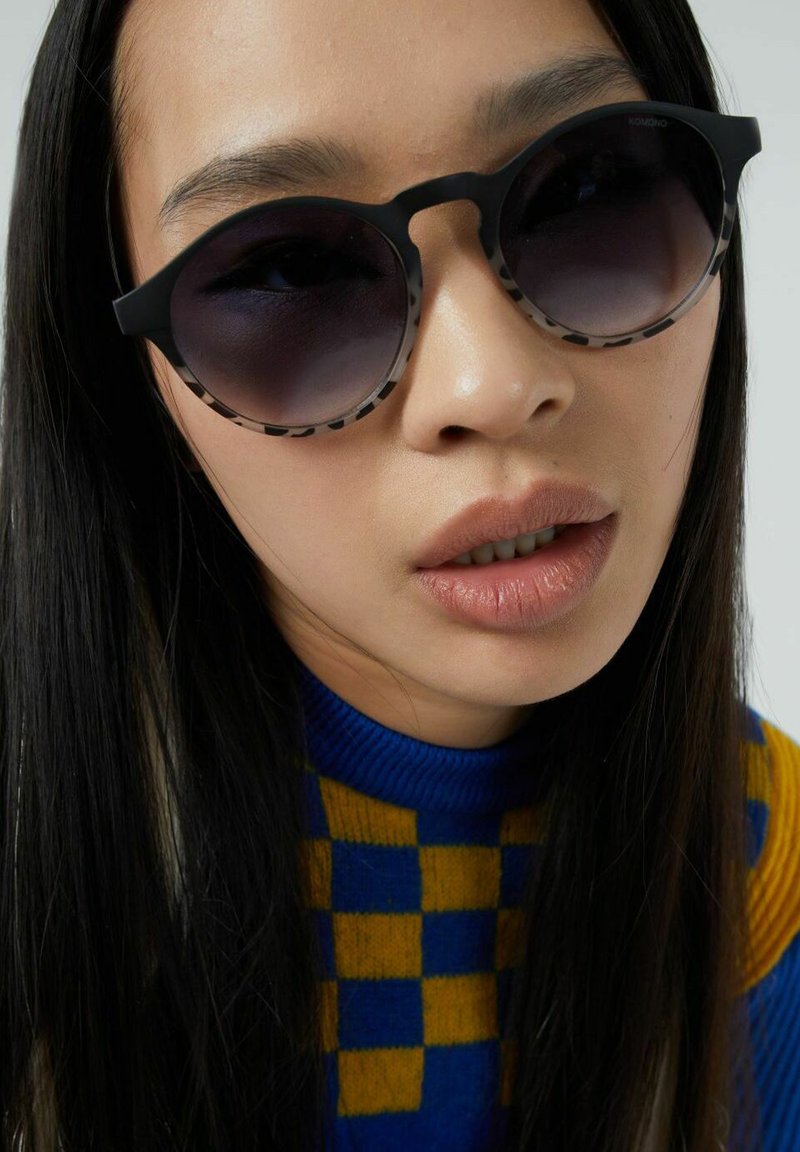 Gafas de sol redondas con montura negra, lentes oscuros y un borde estampado. El modelo lleva un suéter de patrón azul y amarillo. Sin detalles de fondo.