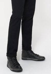 Czarne wysokie trampki z teksturowanego materiału, sznurowanym designem, gumową podeszwą i bocznym zamkiem błyskawicznym, zestawione z podwiniętymi czarnymi spodniami.