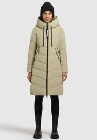 Geacă lungă puffer khaki, cu glugă, quilting în formă de zigzag, fermoar frontal și buzunare laterale, asortată cu colanți negri și cizme.