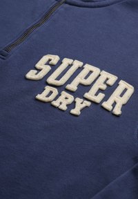 Granatowa bluza z zamkiem błyskawicznym, z teksturowanym kremowym napisem "SUPER DRY" wyraźnie umieszczonym z przodu. Miękki materiał, casualowy fason.
