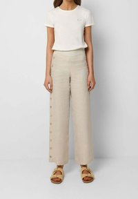 Kvinna som bär en vit kortärmad topp, beiga högmidjade vida byxor med sidoknappar och beiga sandaler med spänne.