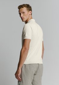 Crème gebreide poloshirt met korte mouwen en een geribbelde kraag, gecombineerd met lichtgrijze linnen shorts. De stof heeft een zachte textuur.