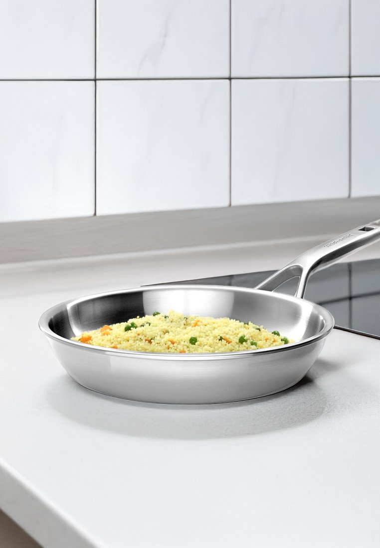 Edelstahlpfanne mit einer glatten, glänzenden Oberfläche, gefüllt mit gelbem Couscous und grünen Erbsen, auf einer weißen Arbeitsplatte platziert.