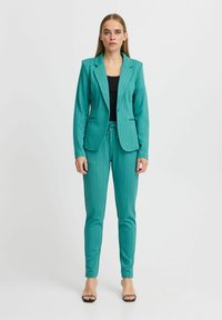 Blazer stampato in teal e pantaloni slim-fit coordinati con vita regolabile. Top nero sotto. Tacchi neri aperti.