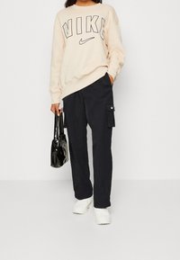 Béžová mikina Nike s výrazným logem, černé cargo kalhoty s kapsou, černá kabelka a bílé tenisky. Neformální sportovní outfit.