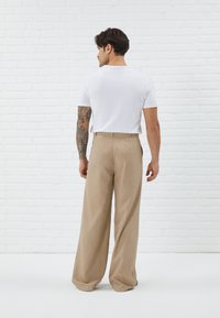 Pantalon large beige avec un motif à carreaux subtil, associé à un t-shirt blanc ajusté. Mannequin de dos devant un mur en briques blanches.