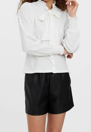 Femme portant un chemisier blanc à manches longues avec un nœud au col et un short noir taille haute sur un fond uni.