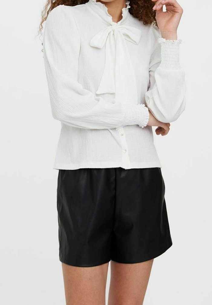 Femme portant un chemisier blanc à manches longues avec un nœud au col et un short noir taille haute sur un fond uni.
