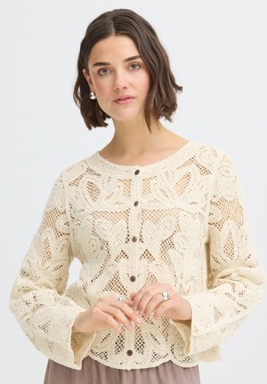 Žena s krátkymi hnedými vlasmi, ktorá nosí krémový háčkovaný sveter na gombíky a strieborné náušnice a prstene, pózuje na bielom pozadí.