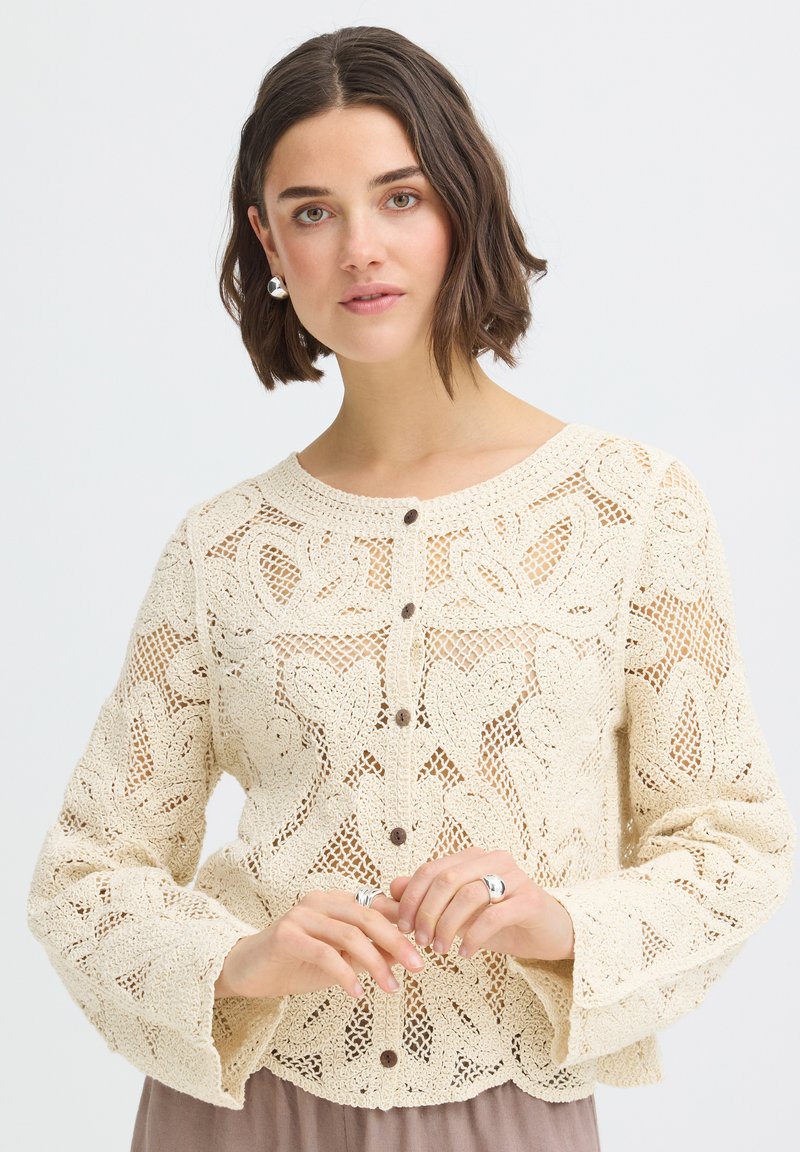 Femme aux cheveux courts bruns portant un pull en crochet crème boutonné, des boucles d'oreilles et des bagues en argent, posant sur fond blanc.
