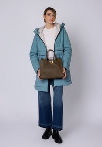 Brązowa torebka z teksturowanym wykończeniem, złotymi dodatkami i podwójnymi uchwytami. Modelka nosi niebieską kurtkę puchową i szerokie dżinsy, stojąc na jednolitym tle.