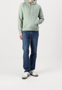 Světlounce zelená mikina s kapucí a přední kapsou s bílým logem, v kombinaci s modrými džínami a šedobílými sportovními botami s černými pruhy.