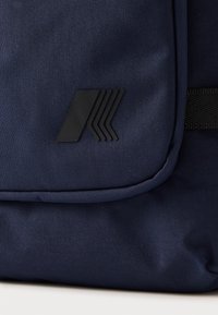 Tmavomodrý látkový batoh s hladkou textúrou, predným klapkou a čiernym logom s geometrickými líniami.