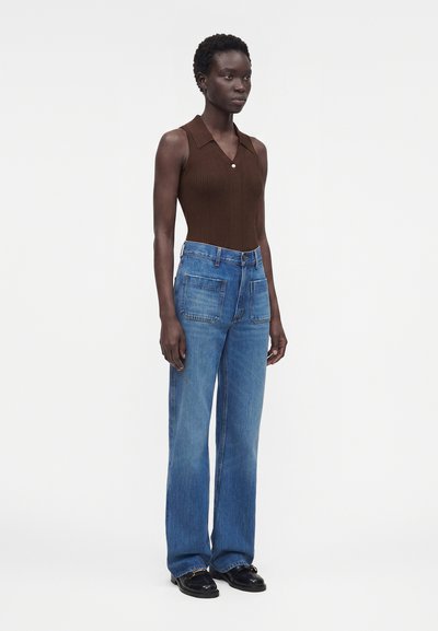Γυναίκα στέκεται φορώντας ένα καφέ πλεκτό αμάνικο τοπ, μπλε ψηλόμεσα φαρδιά τζιν με μεγάλες μπροστινές τσέπες και μαύρα loafers.