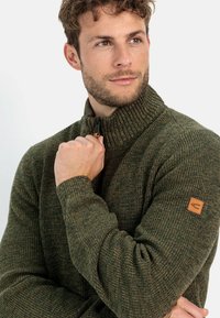Strick-Jacke mit Reißverschluss in dunkelgrün, mit strukturiertem Muster, geripptem Kragen und Manschetten, versehen mit einem Leder-Logopatch auf dem linken Ärmel.