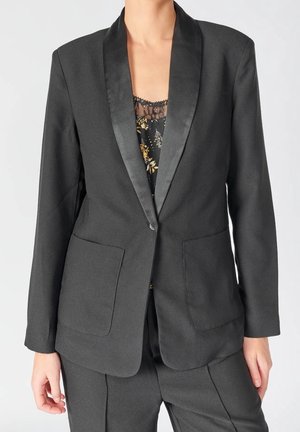 Frau trägt eine maßgeschneiderte schwarze Anzugjacke mit Satinrevers über einem floralen, spitzenbesetzten Top, kombiniert mit passenden schwarzen Hosen.
