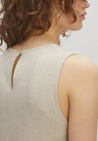 Beige mouwloze jurk met een textuurstof, een sleutelgatuitsnijding op de rug en een kleine knoopsluiting bij de halslijn.