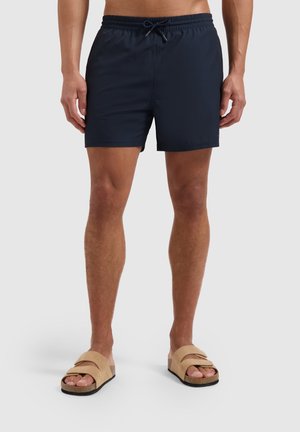Marineblaue Badeshorts mit elastischem Bund und Kordelzug, aus leichtem Material. Kombiniert mit sandfarbenen Slidesandalen.