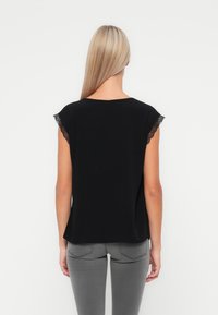 Kvinna med långt blont hår som bär en svart ärmlös topp med spetskant och grå jeans, stående mot en enfärgad vit bakgrund.