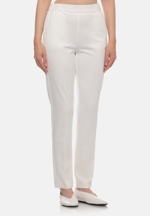 Donna che indossa pantaloni bianchi a gamba dritta con vita elastica e scarpe basse bianche appuntite, in piedi davanti a uno sfondo semplice.