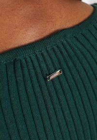 Primo piano di una spalla di pelle scura che indossa un top a coste verde con un piccolo tag metallico che legge "SLOGAN" vicino al collo.