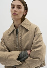 Beige, kort jacka med en stor krage, dragkedjestängning och voluminösa ärmar. Bärs över en texturerad grå tröja. Slät, matt yta.