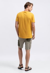 T-shirt jaune en coton, col rond. Shorts gris clair avec poches latérales vert olive. Porté avec des sandales noires, vue de dos.