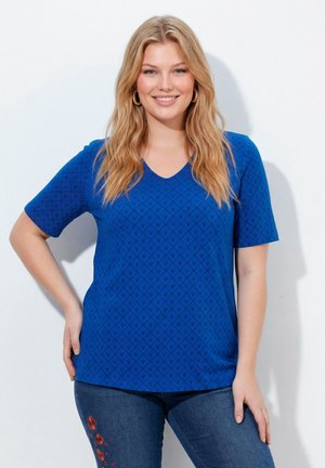 Lächelnde Frau mit langen blonden Haaren trägt ein blau gemustertes kurzärmeliges Hemd und Jeans mit rotem Blumendesign am Oberschenkel, steht vor einer weißen Wand.