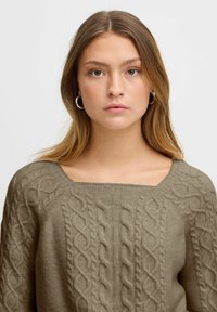 Grüner, kabelgestrickter Pullover mit quadratischem Ausschnitt, strukturiertem Muster und weichem Stoff. Betont durch Details an den Ärmeln und gerippte Kanten.