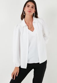 White button-up blouse with puffed sleeves, smooth fabric, and a pointed collar, layered over a white V-neck top, paired with black pants.
